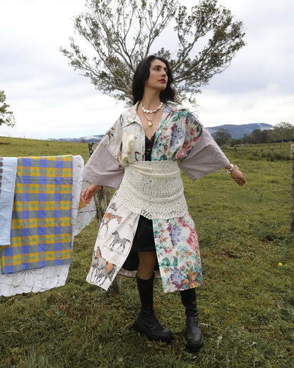 A woman stands outdoors in a grassy field wearing a bohemian-style patchwork dress with floral and animal prints, a white crochet waist apron, and black boots. A blue and yellow plaid blanket is hanging on a line to the left, with trees and hills in the background.