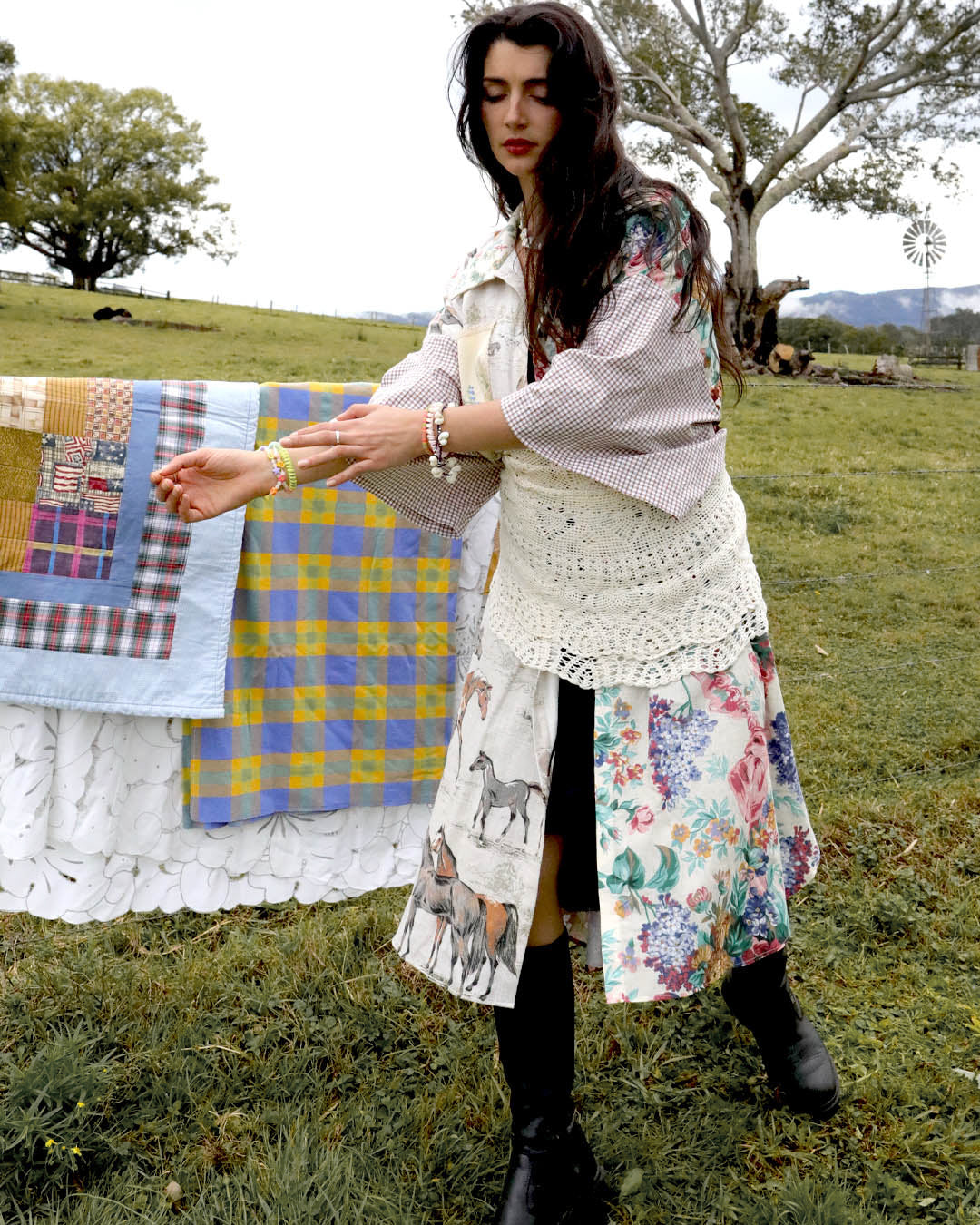 A woman stands outdoors in a grassy field wearing a long bohemian-style patchwork dress with floral and animal prints, layered with a white crochet vest and striped shirt. A colorful quilt is draped over a stand beside her.