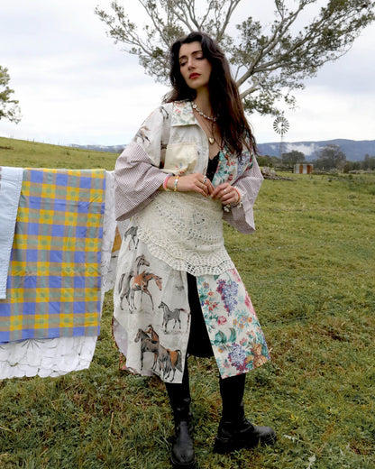 A woman stands in a grassy outdoor field wearing a long, bohemian-style printed coat with horse and floral motifs, layered with a cream crochet apron and black boots. A blue and yellow plaid blanket is draped over a tree to her left, with hills and a windmill in the background.