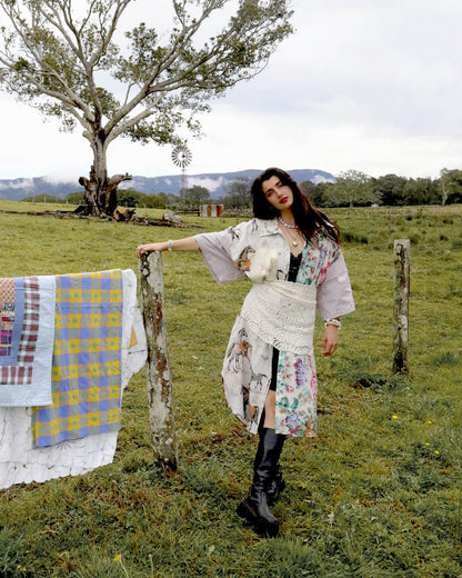 A woman stands in a grassy field wearing a long-sleeved, asymmetrical bohemian-style dress with a floral and abstract print, a white crochet overlay, and black knee-high boots. She is posed next to a rustic wooden fence post with a quilt hanging on it, with a large tree and mountains in the background.