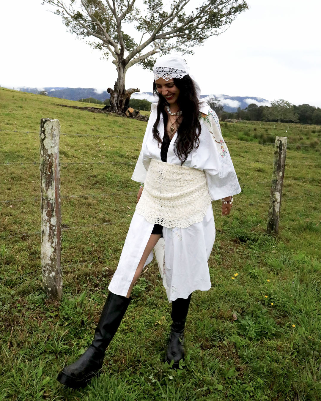 A woman stands in a grassy outdoor field wearing a white bohemian-style outfit with a long-sleeved top, a textured wrap skirt, a white headpiece, and black boots.