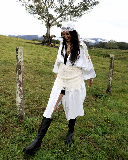 A woman stands in a grassy outdoor field wearing a white bohemian-style outfit with a long-sleeved top, a textured wrap skirt, a white headpiece, and black boots.