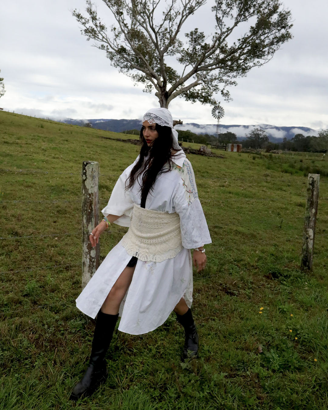 A woman wearing a white Dear Abigael oversized shirt dress made from heirloom patchwork fabrics with a matching headscarf, cream waist sash, and black knee-high boots, posing in a grassy outdoor field with a tree and hills in the background.