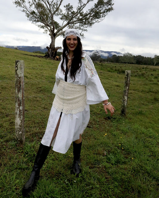A woman wearing a white patchwork heirloom Dear Abigael shirt dress with a cream lace waist apron, black knee-high boots, and a white headband stands outdoors in a grassy field with a tree and hills in the background.