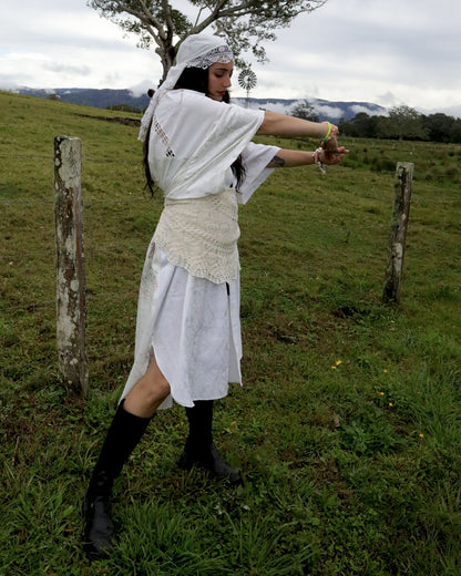 A woman wearing a white dress with a textured lace waist sash, black knee-high boots, and a white headscarf poses in a grassy outdoor field under an overcast sky.