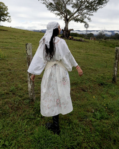 A woman stands in a grassy field wearing a white embroidered patchwork vintage dress with a matching head covering and black boots, photographed outdoors with hills, trees, and an overcast sky in the background.