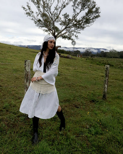 A woman stands in a grassy outdoor field wearing a white long-sleeve dress with a cream lace-trimmed waist in Copenhagen fashion week style, black knee-high socks, and a white headband. A tree, fence posts, and distant hills are visible in the background under an overcast sky.