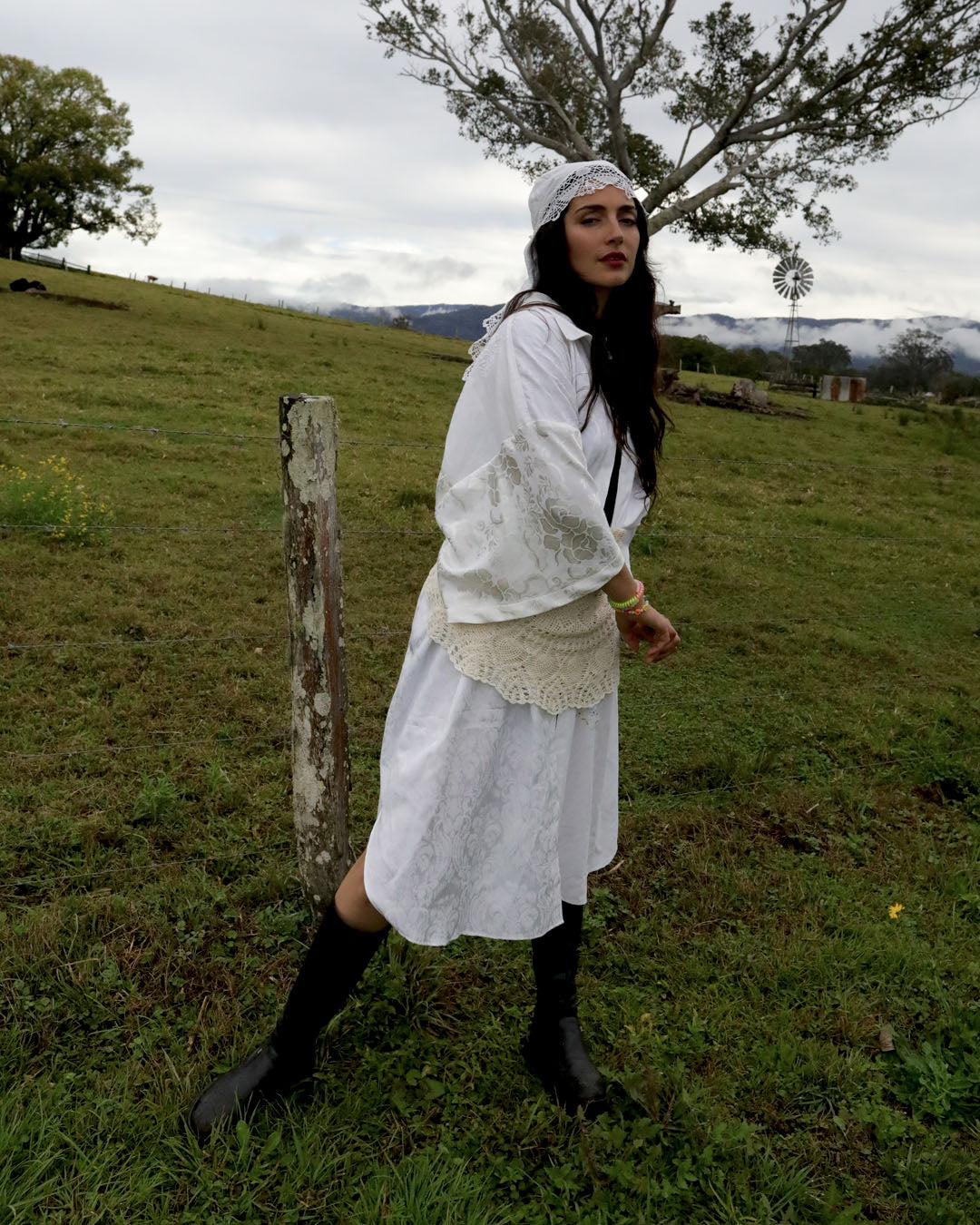 A woman wearing a white lace dress, white headband, and black knee-high boots stands in a grassy outdoor field with trees and hills in the background.