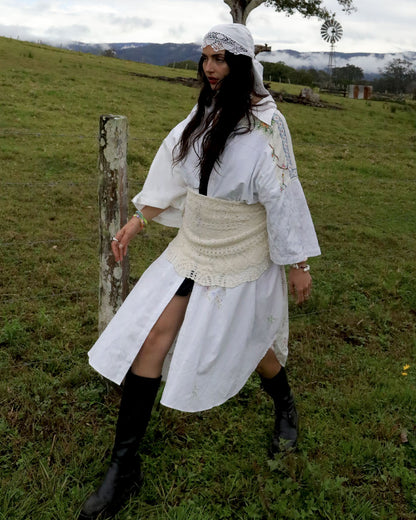 A woman wearing a white Dear Abigael shirt dress with a cream lace corset, white lace headband, and black knee-high boots, standing in a grassy field.