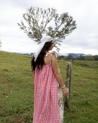 A woman stands outdoors in a grassy field wearing a red and white gingham dress with a white lace headscarf, seen from behind.