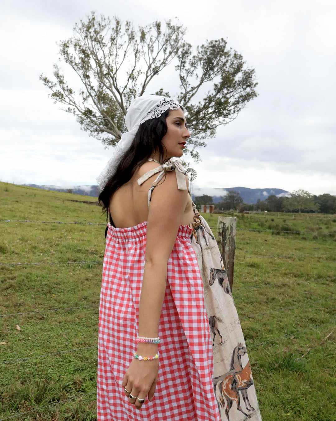 A woman stands in a grassy field wearing a red and white gingham dress with a white lace head covering and a horse-patterned shawl draped over her arm. Trees and hills are visible in the background under an overcast sky.