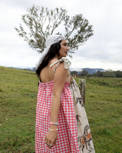 A woman stands in a grassy field wearing a red and white gingham dress with a white lace head covering and a horse-patterned shawl draped over her arm. Trees and hills are visible in the background under an overcast sky.