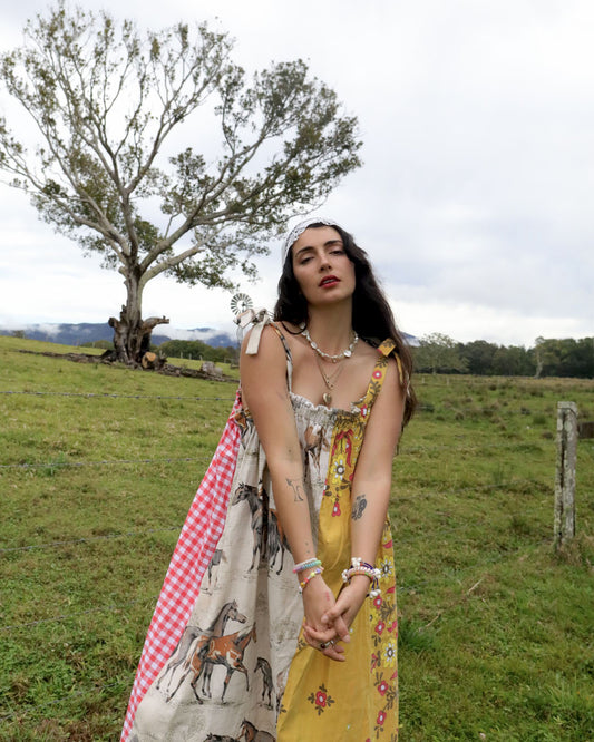 A woman stands in a grassy outdoor field wearing a bohemian-style dress with a red-and-white gingham panel, a horse-print section, and a yellow floral section. She is accessorized with layered necklaces and bracelets, with a tree and open landscape in the background.