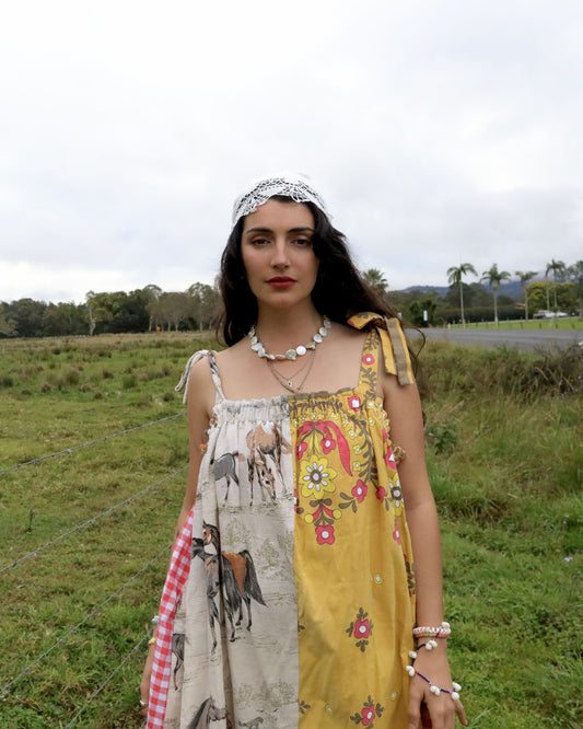 A woman stands outdoors in a grassy field wearing a sleeveless patchwork dress with floral and animal prints, layered necklaces, bracelets, and a white headband.