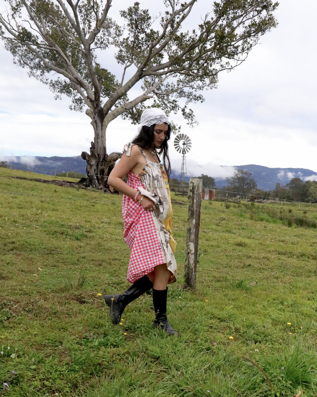 A woman stands in a grassy outdoor field wearing a distressed, multi-colored dress with red and white gingham, a floral print, and yellow panels, paired with black boots and a white headscarf.