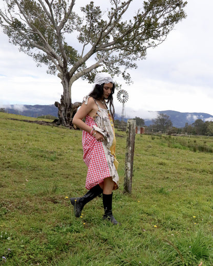 A woman stands in a grassy outdoor field wearing a distressed, multi-colored dress with red and white gingham, a floral print, and yellow panels, paired with black boots and a white headscarf.