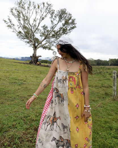 A woman wearing a bohemian-style patchwork maxi dress with horse prints, a red gingham panel, and a yellow floral side, standing in a grassy field with a tree in the background.