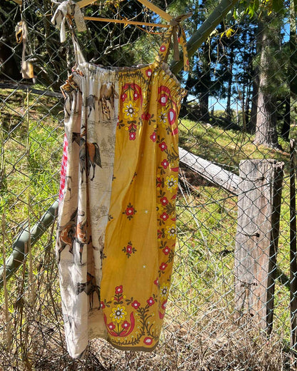 A sleeveless two-tone dress ohanging outdoors on a fence in a grassy wooded area. The fabric is split vertically: one side is cream with a horse and rider print, and the other side is mustard yellow with red and pink floral embroidery and motifs.