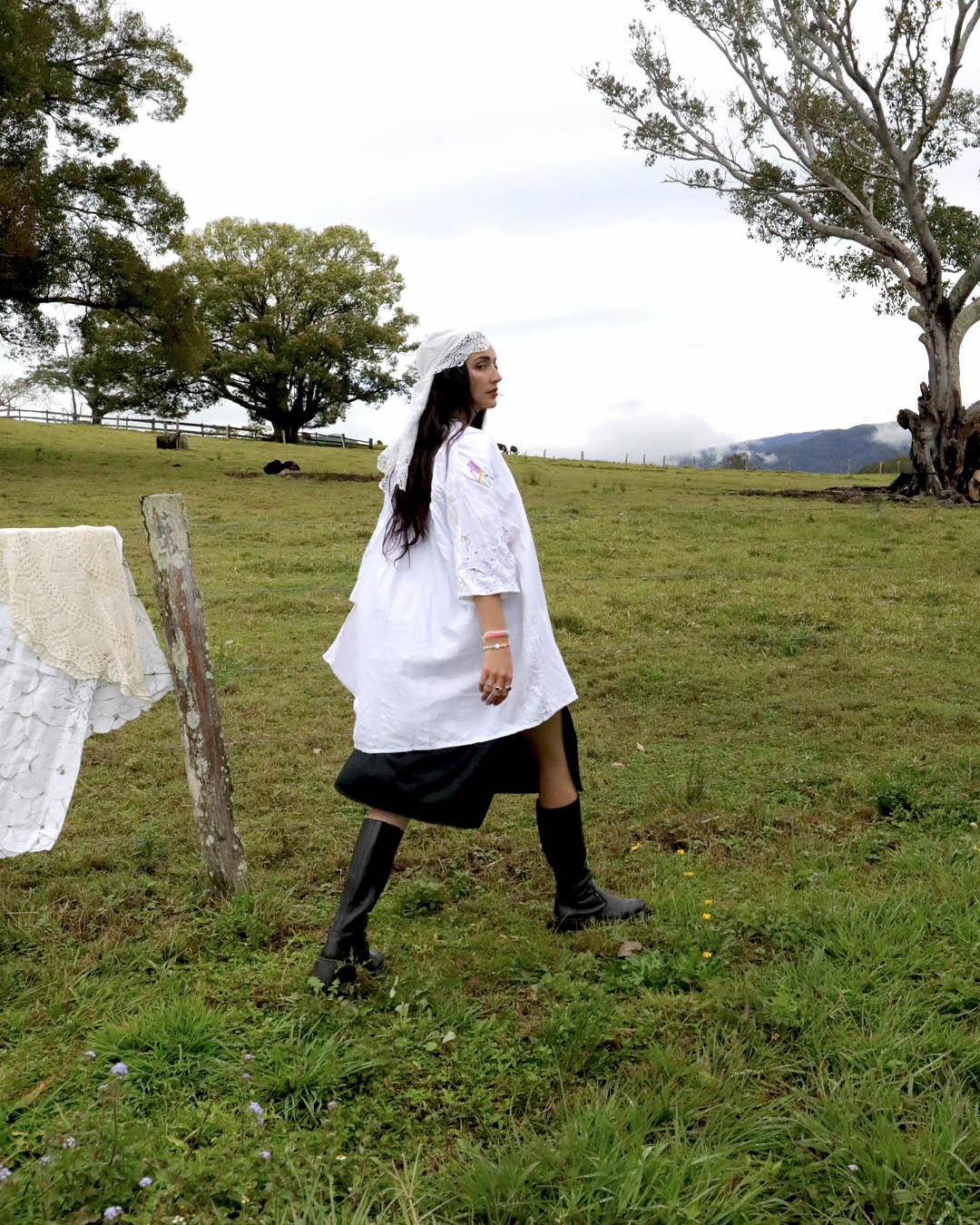A woman walking across a grassy field wearing a white lace-trimmed Dear Abigael heirloom smock shirt dress with a black skirt and black knee-high boots. Trees and a cloudy sky are visible in the background.