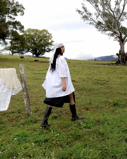 A woman walking across a grassy field wearing a white lace-trimmed Dear Abigael heirloom smock shirt dress with a black skirt and black knee-high boots. Trees and a cloudy sky are visible in the background.