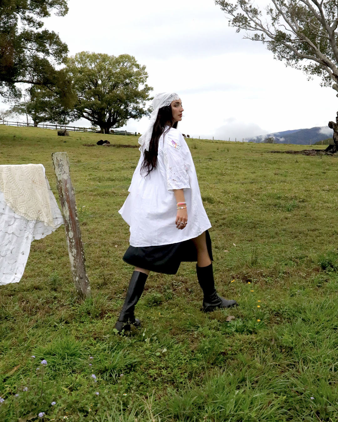A woman walks across a grassy field wearing a white lace-trimmed tunic dress with a black skirt, black knee-high socks, and black boots. She has a white veil on.