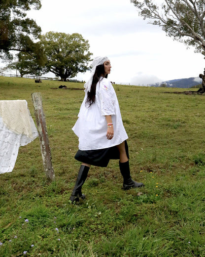 A woman walks across a grassy field wearing a white lace-trimmed tunic dress with a black skirt, black knee-high socks, and black boots. She has a white veil on.
