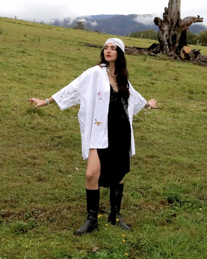 A woman stands in a grassy outdoor field wearing a white embroidered open-front shirt over a black dress, with black knee-high boots and a white headband. Mountains and a tree are visible in the background.