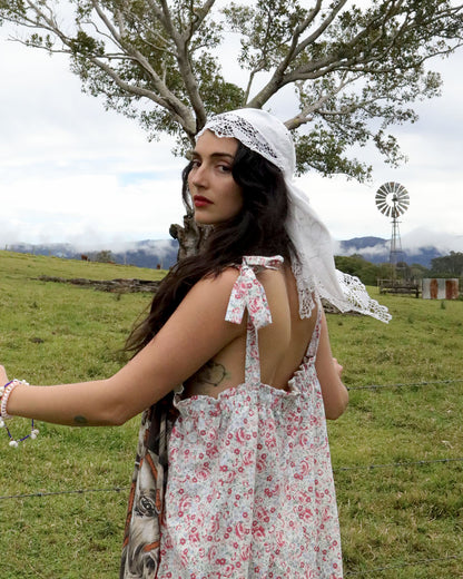 A woman wearing a bohemian-style floral dress and white lace headscarf stands in a grassy field with a tree and windmill in the background.