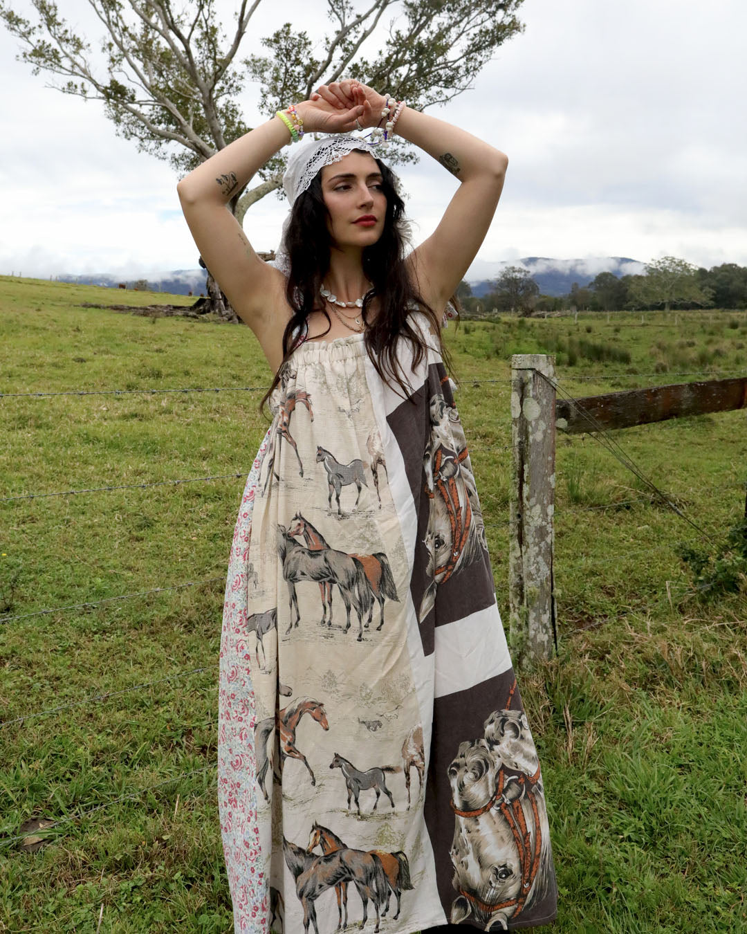 A woman stands outdoors in a grassy field wearing a long patchwork dress with horse illustrations. The dress has a cream base with brown, gray, and floral patterned panels.