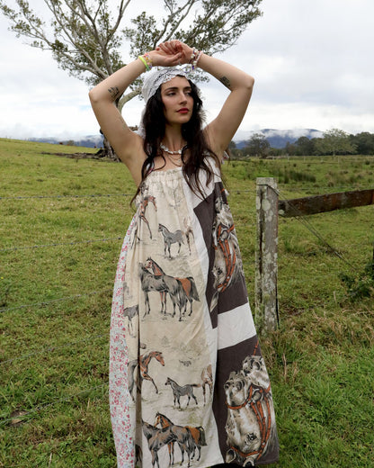 A woman stands outdoors in a grassy field wearing a long patchwork dress with horse illustrations. The dress has a cream base with brown, gray, and floral patterned panels.