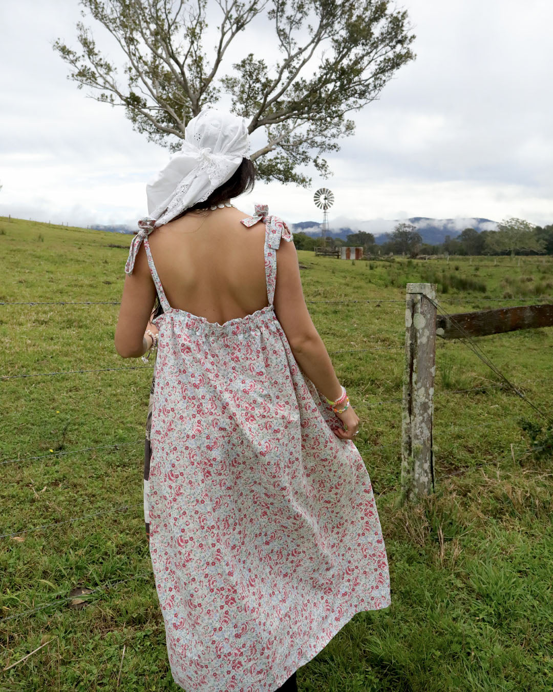A woman viewed from behind wearing a white bonnet and a sleeveless backless dress with a red and green floral print, standing in a grassy field.