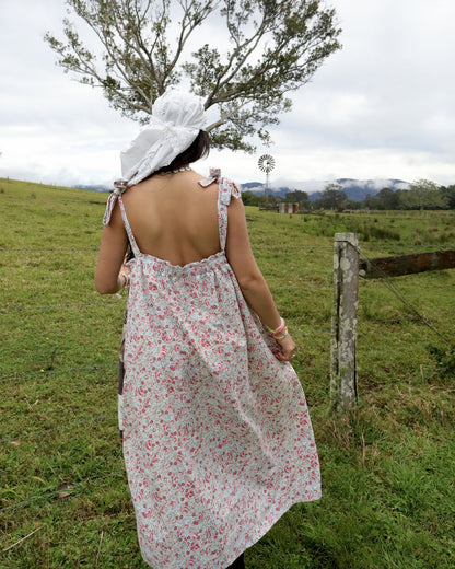 A woman viewed from behind wearing a white bonnet and a sleeveless backless dress with a red and green floral print, standing in a grassy field.