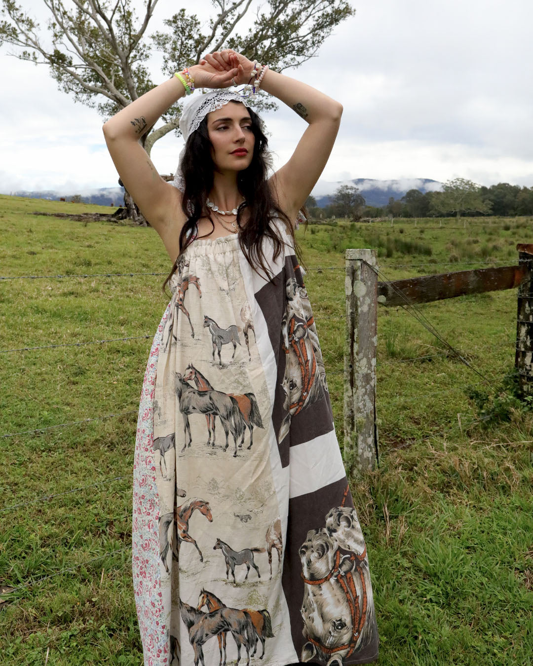 A woman stands outdoors in a grassy field wearing a sleeveless bohemian-style maxi dress with a horse print. The dress features a cream base with brown, black, and floral patterns.