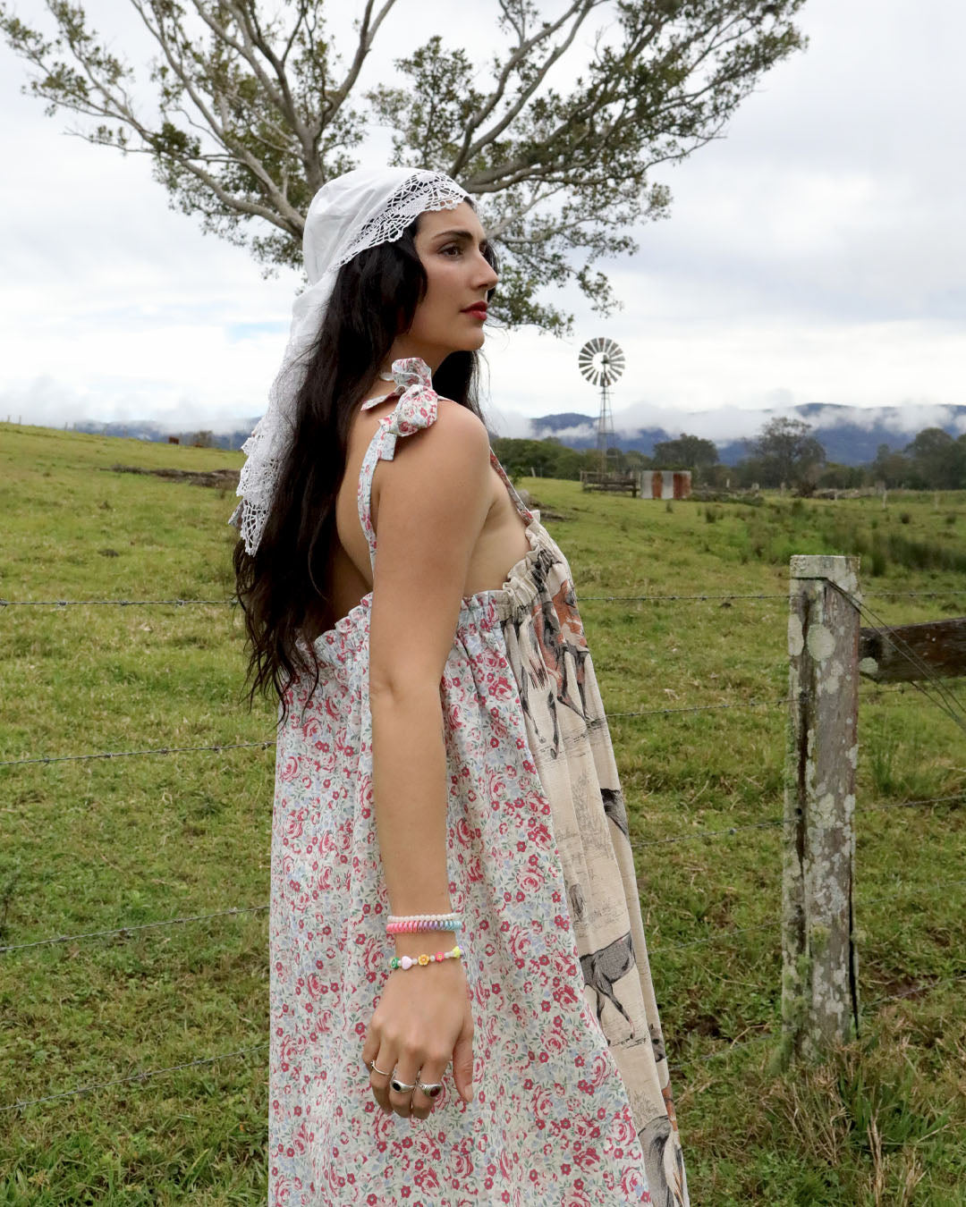 A woman wearing a floral sleeveless dress and a white lace-trimmed headscarf stands in profile in an outdoor grassy field with a fence, tree, and windmill in the background.