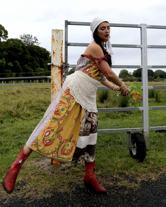 A woman wearing a Dear Abigael one size adjustable smock dress using patchwork vintage fabrics. One is mustard and one is horse print. She is kicking one leg out while holding nasturtiums in front of a farm gate