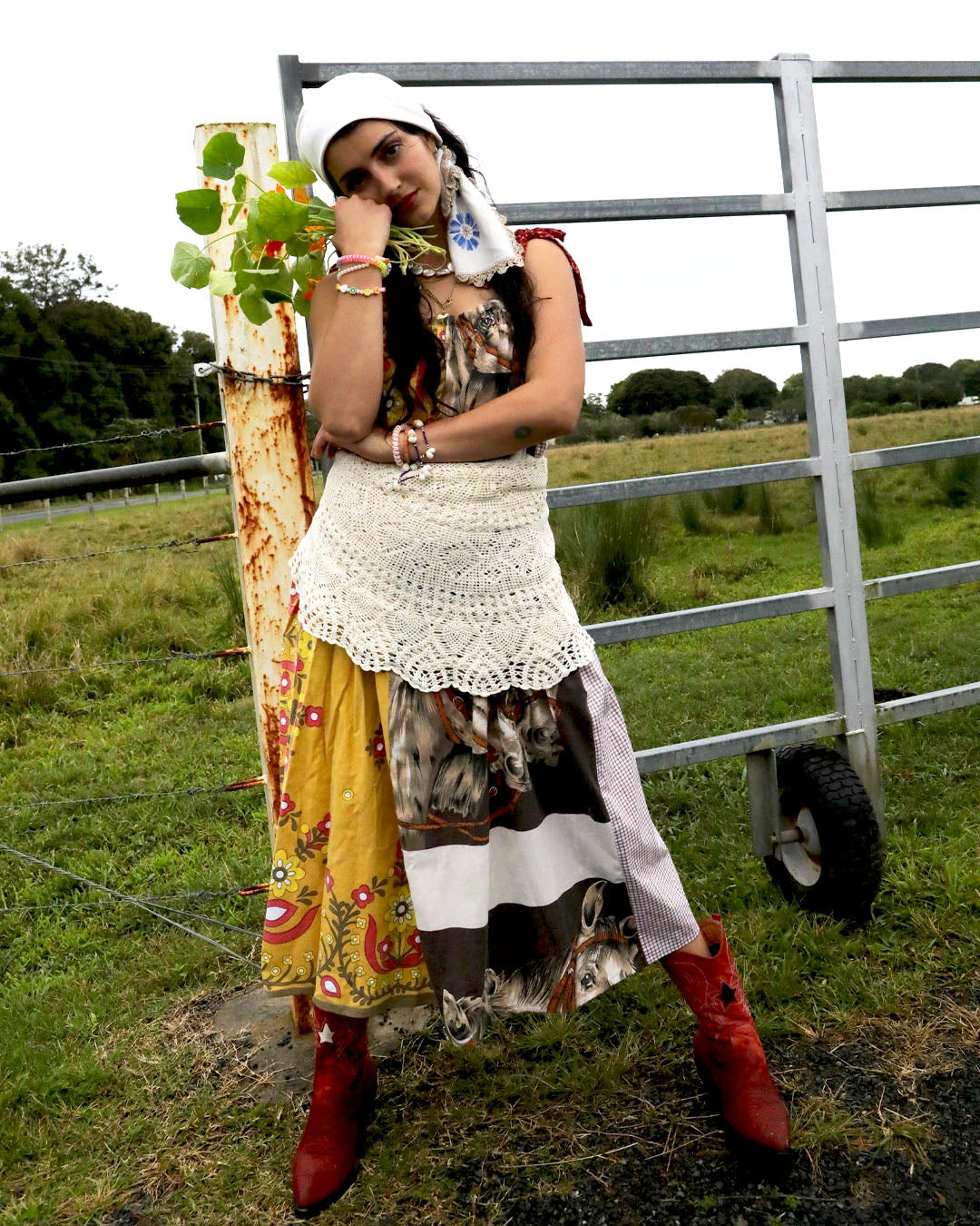 A woman wearing a smock dress made from vintage patchwork fabrics and a crochet apron with layered necklaces and bracelets and an embroidered headscarf. She is holding nasturtiums and wearing red cowboy boots and standing in front of a farm gate.