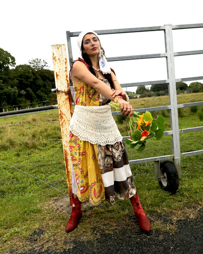 A woman wearing a smock dress made from vintage patchwork fabrics and a crochet apron with layered necklaces and bracelets and an embroidered headscarf. She is holding nasturtiums and wearing red cowboy boots and standing in front of a farm gate.