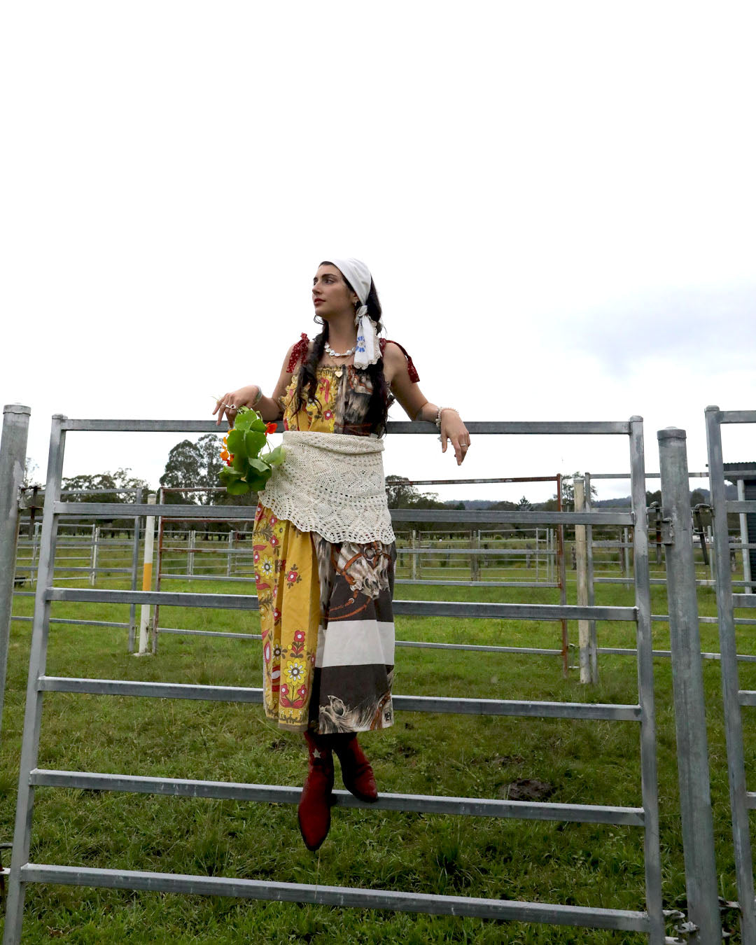 A woman wearing a smock dress made from vintage patchwork fabrics and a crochet apron with layered necklaces and bracelets and an embroidered headscarf. She is holding nasturtiums and wearing red cowboy boots and standing up on a farm gate.