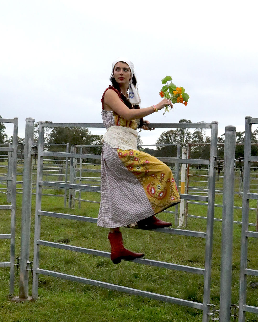 A woman wearing a smock dress made from vintage patchwork fabrics and a crochet apron with layered necklaces and bracelets and an embroidered headscarf. She is holding nasturtiums and wearing red cowboy boots and climbing  a farm gate.