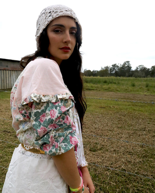 A woman wearing a vintage-style floral blouse with ruffled sleeves, a white lace headband, and a white skirt, standing in an outdoor field.