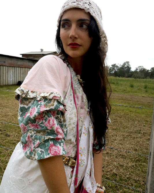 A woman wearing a vintage-style bohemian outfit with a pink and green floral puff-sleeve top, white dress, and lace headband, standing outdoors in a grassy field.