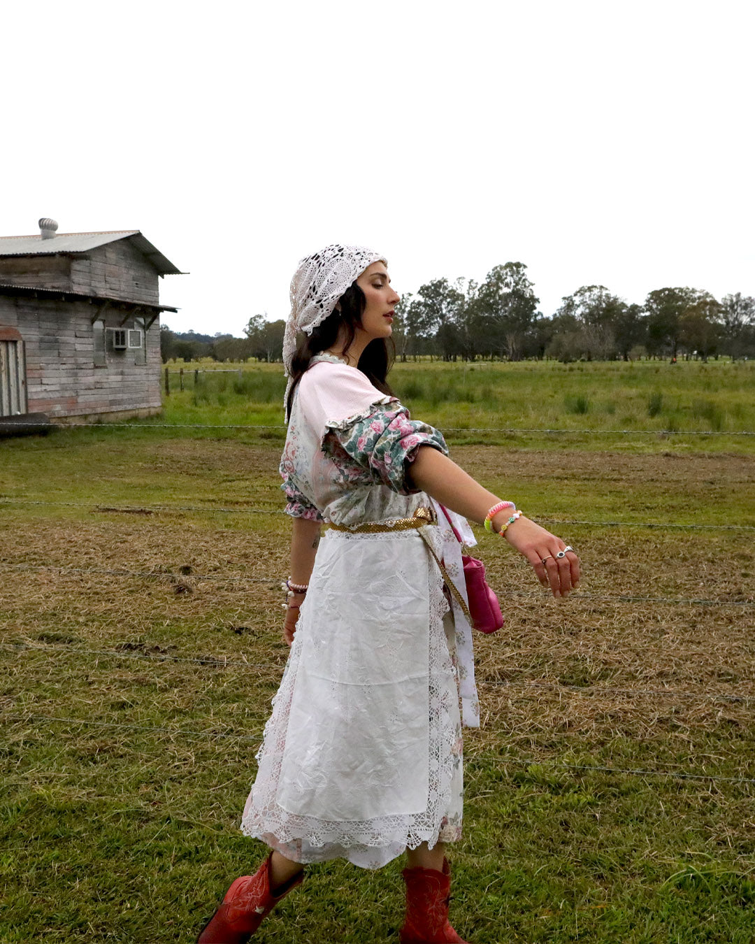 A woman wearing a white embroidered dress, floral long-sleeve top, white headscarf, and red boots walks across a grassy field with a rustic wooden building in the background.