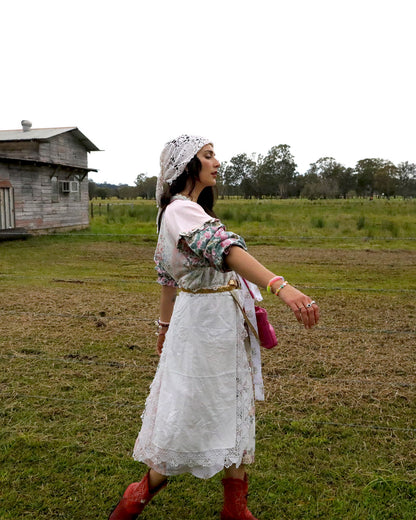 A woman wearing a white embroidered dress, floral long-sleeve top, white headscarf, and red boots walks across a grassy field with a rustic wooden building in the background.