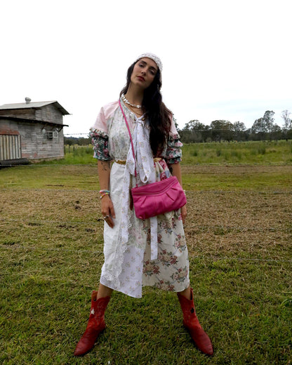 A woman stands outdoors in a grassy field wearing a white floral midi dress with a tie neckline, puff sleeves, a white headband, layered necklaces, a bright pink crossbody bag, and red cowboy boots.