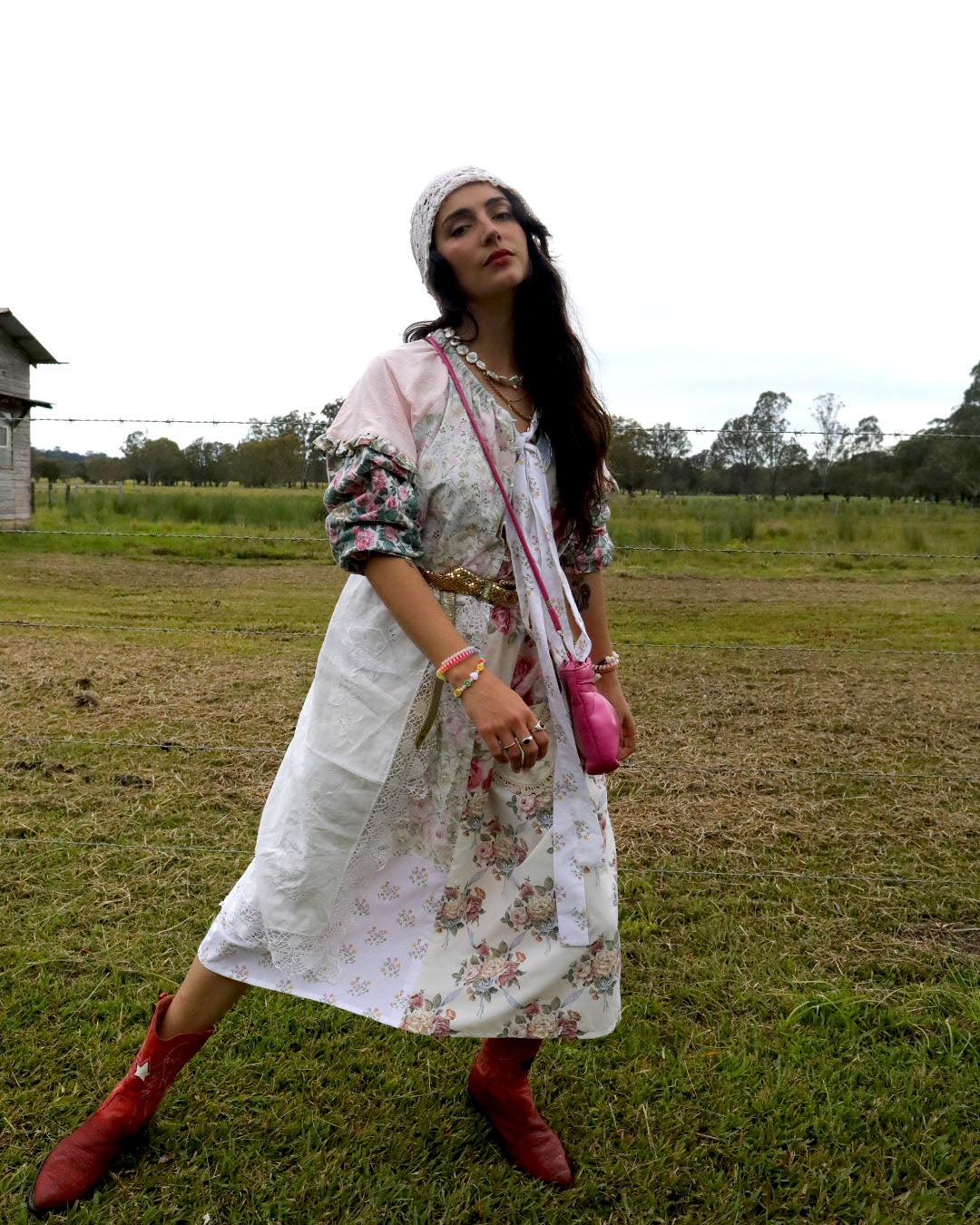 A woman stands in an open grassy field wearing a long white dress with a pink and green floral print, a white headband, layered necklaces, a pink crossbody bag, and red boots.