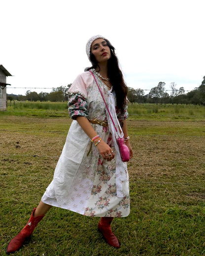 A woman stands in an open grassy field wearing a long white dress with a pink and green floral print, a white headband, layered necklaces, a pink crossbody bag, and red boots.