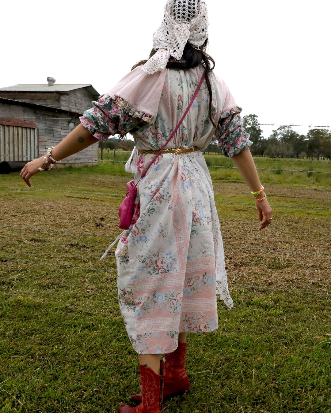 A person standing outdoors in a grassy field wearing a long floral dress with a pink shawl, a white lace head covering, red boots, and carrying a pink crossbody bag.