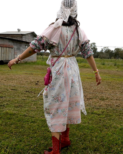 A person standing outdoors in a grassy field wearing a long floral dress with a pink shawl, a white lace head covering, red boots, and carrying a pink crossbody bag.
