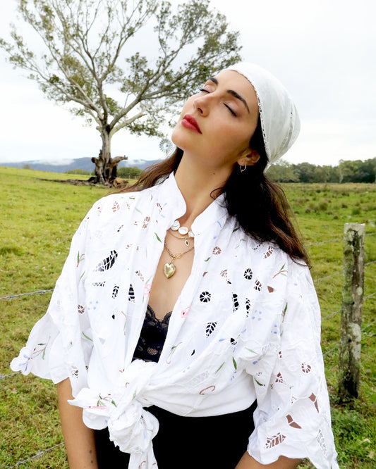 A woman outdoors in a grassy field wearing a white embroidered eyelet shirt tied at the waist, a white headscarf, layered gold necklaces, and a black top.