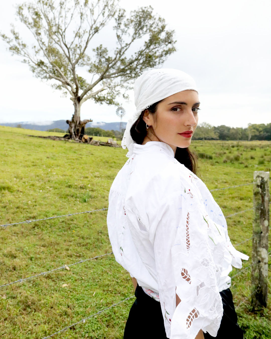 A woman wearing a white embroidered blouse with lace sleeves and a matching white headscarf, standing in a grassy field with a tree and fence post in the background.